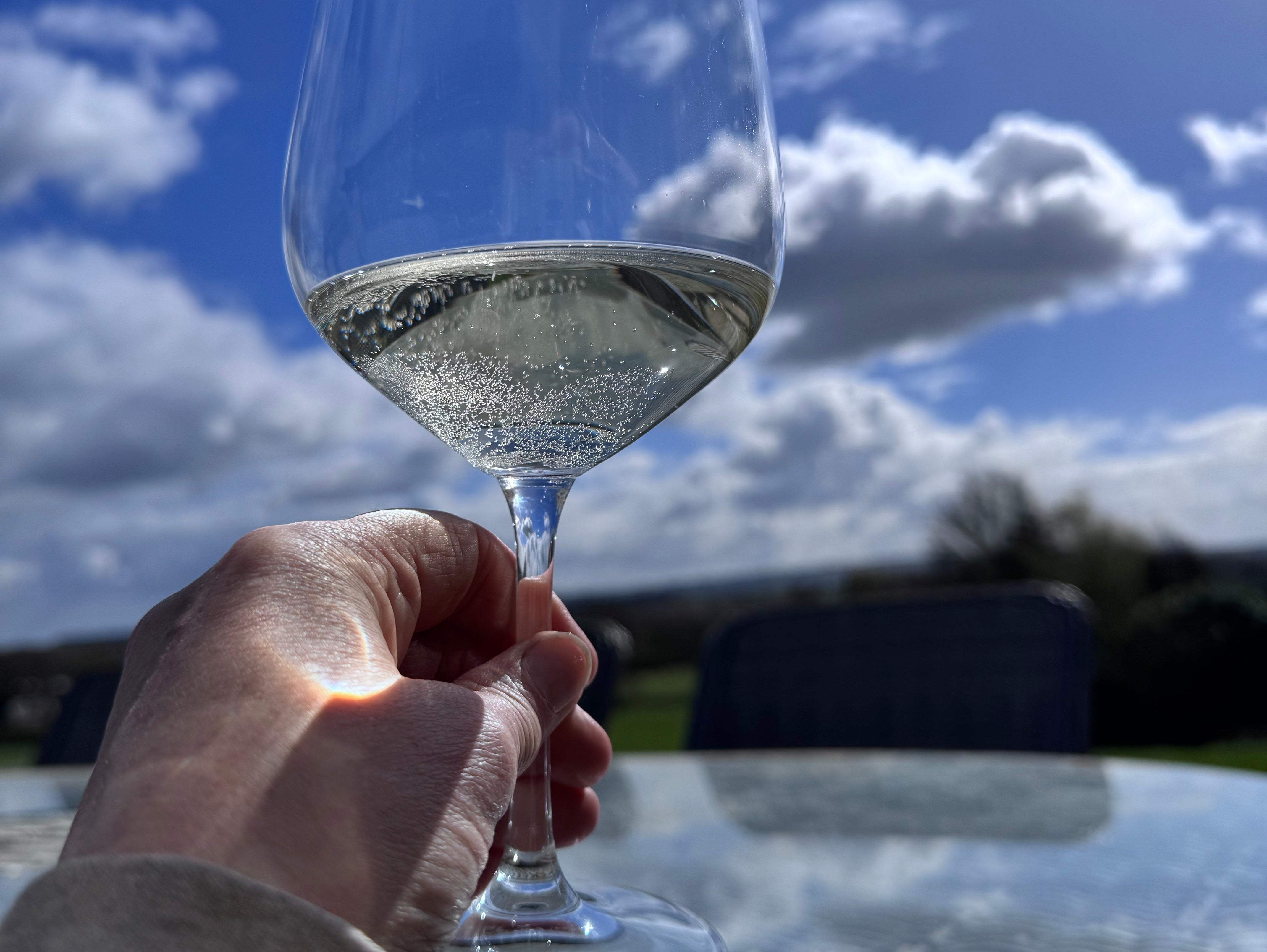 A glass of white wine in a hand against a blue sky with white clouds