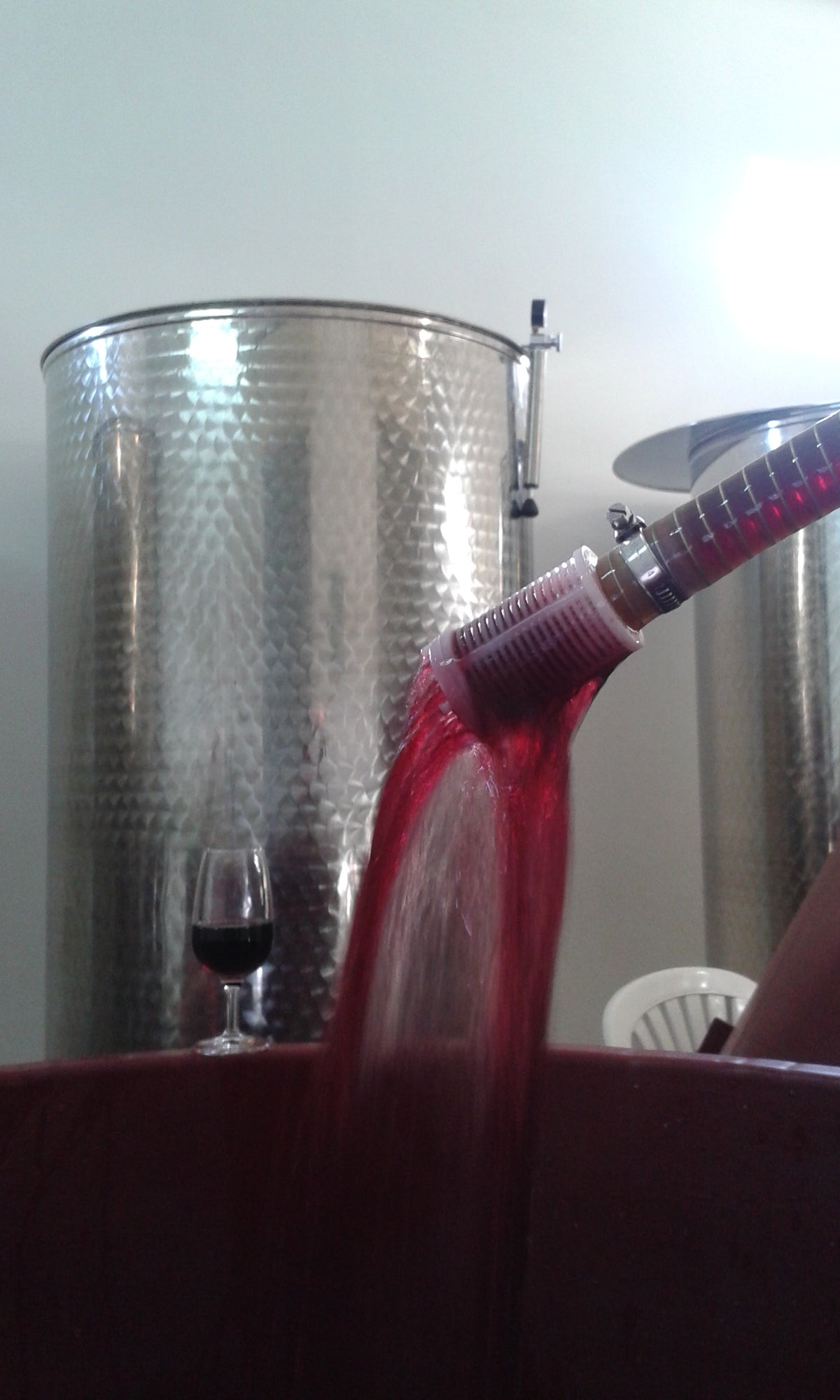 Red wine being poured from a large metal vat into a barrel or cask, with a small wine glass visible in the background.