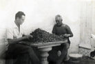 Black and white vintage photograph showing two men seated at a table piled with dark grapes