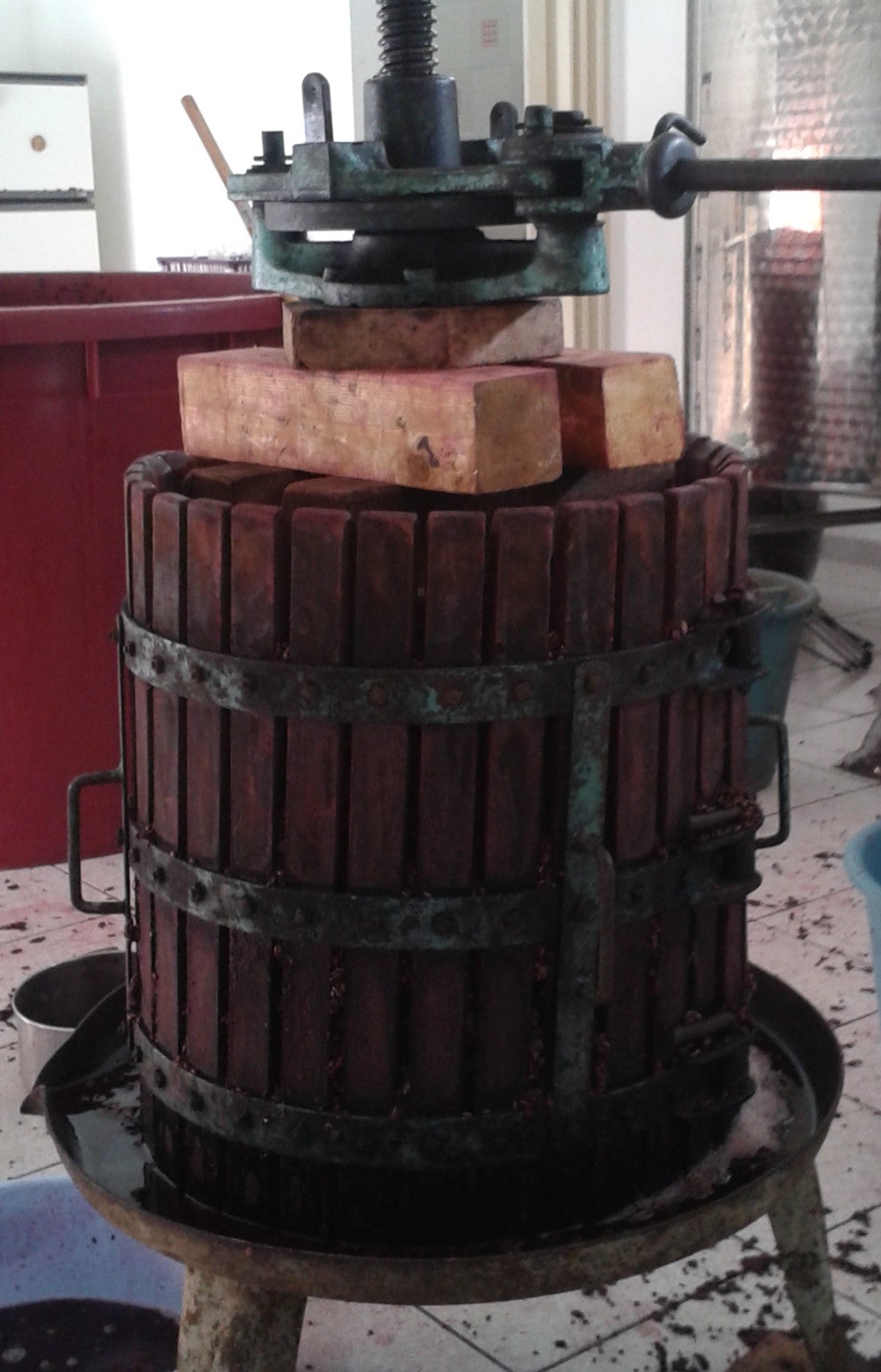 A traditional small wooden grape press with red juice at bottom, with stainless steel tank behind